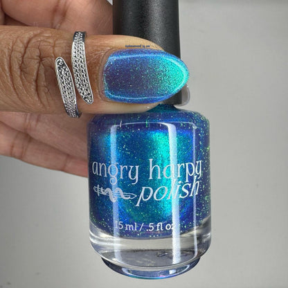 A hand holding a bottle of Angry Harpy Polish nail lacquer, showing the blue and green iridescent glitter polish on both the bottle and a fingernail. The hand is wearing two silver rings.