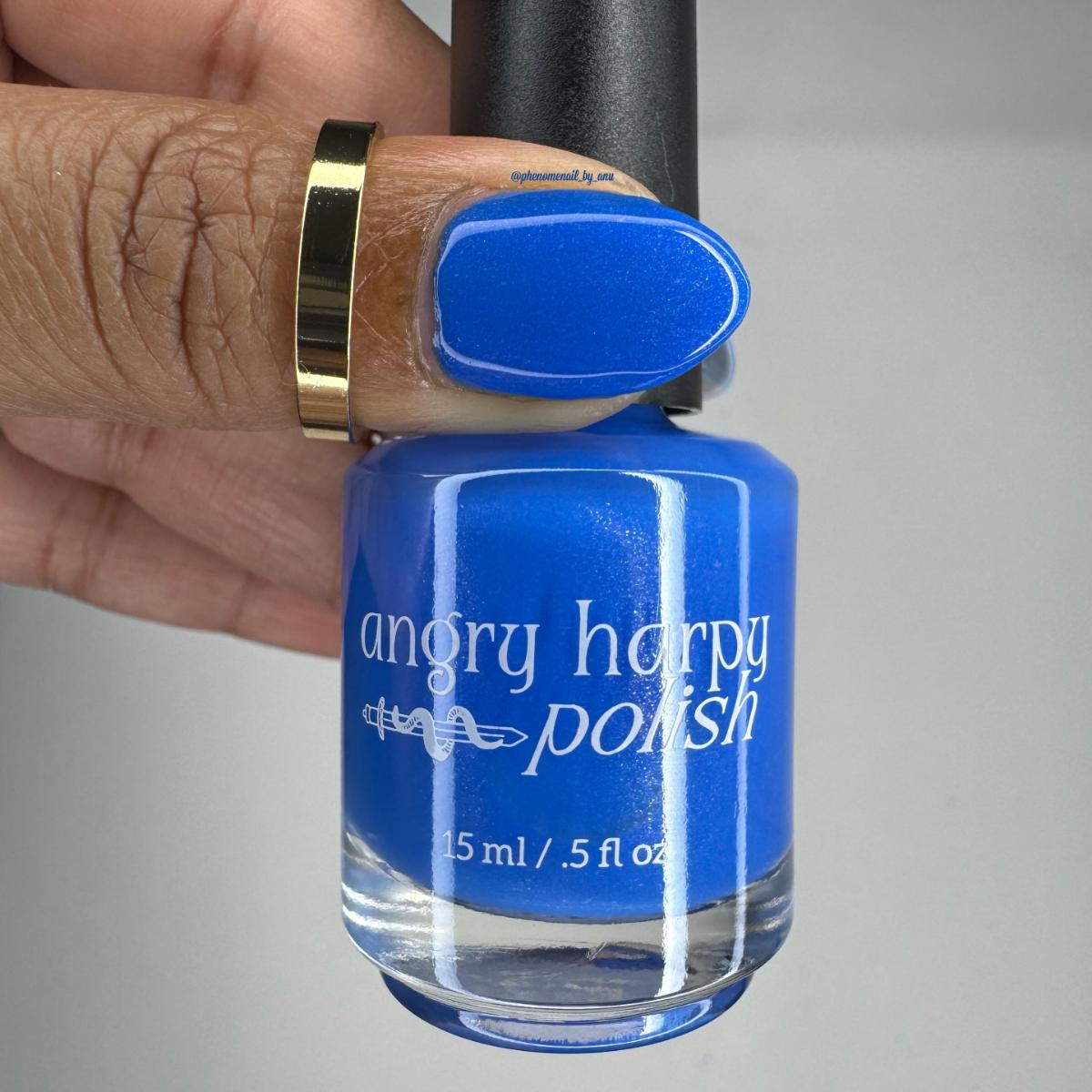 A hand holding a bottle of Angry Harpy Polish nail lacquer, showing the bright royal blue color on the fingernail and in the bottle. The hand is wearing a gold ring, and the background is a plain light gray.