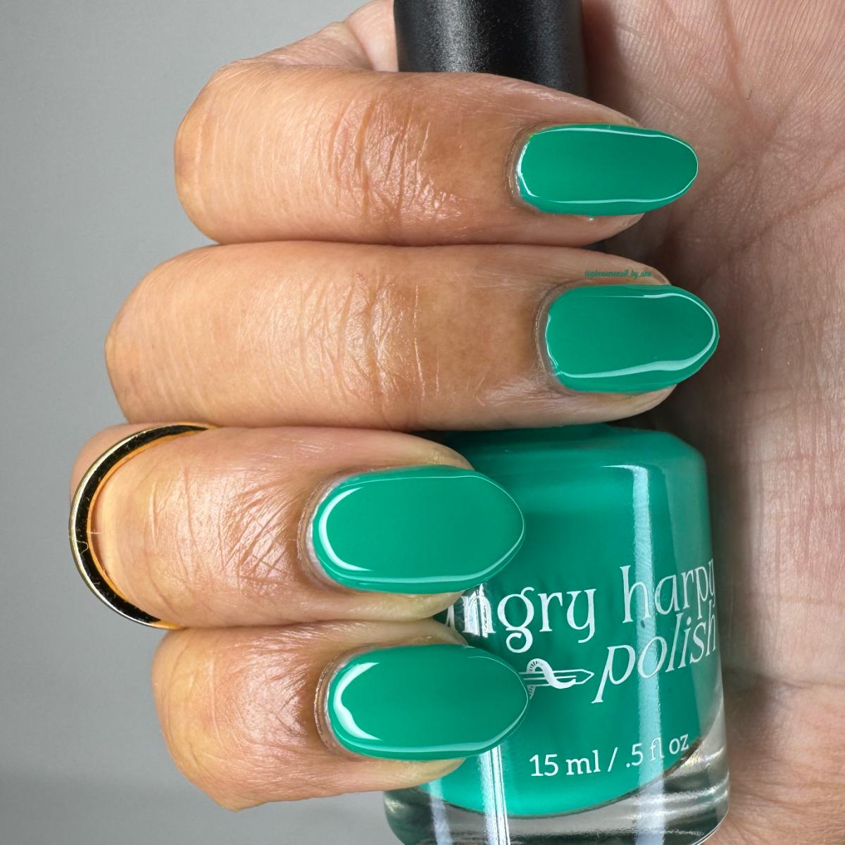 A close-up of a hand holding a bottle of Angry Harp Polish nail lacquer, showing the green polish applied to the fingernails against a neutral gray background.