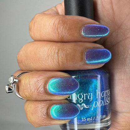 A close-up of a hand holding a bottle of holographic nail polish. The nails are painted in a shimmering blue-to-purple iridescent finish, and the bottle label reads 'angry harpy polish'.