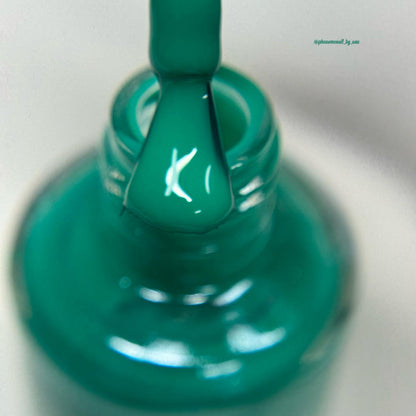 A close-up of a glossy teal green nail polish brush applying polish to a nail inside a matching teal green bottle, set against a soft white background.