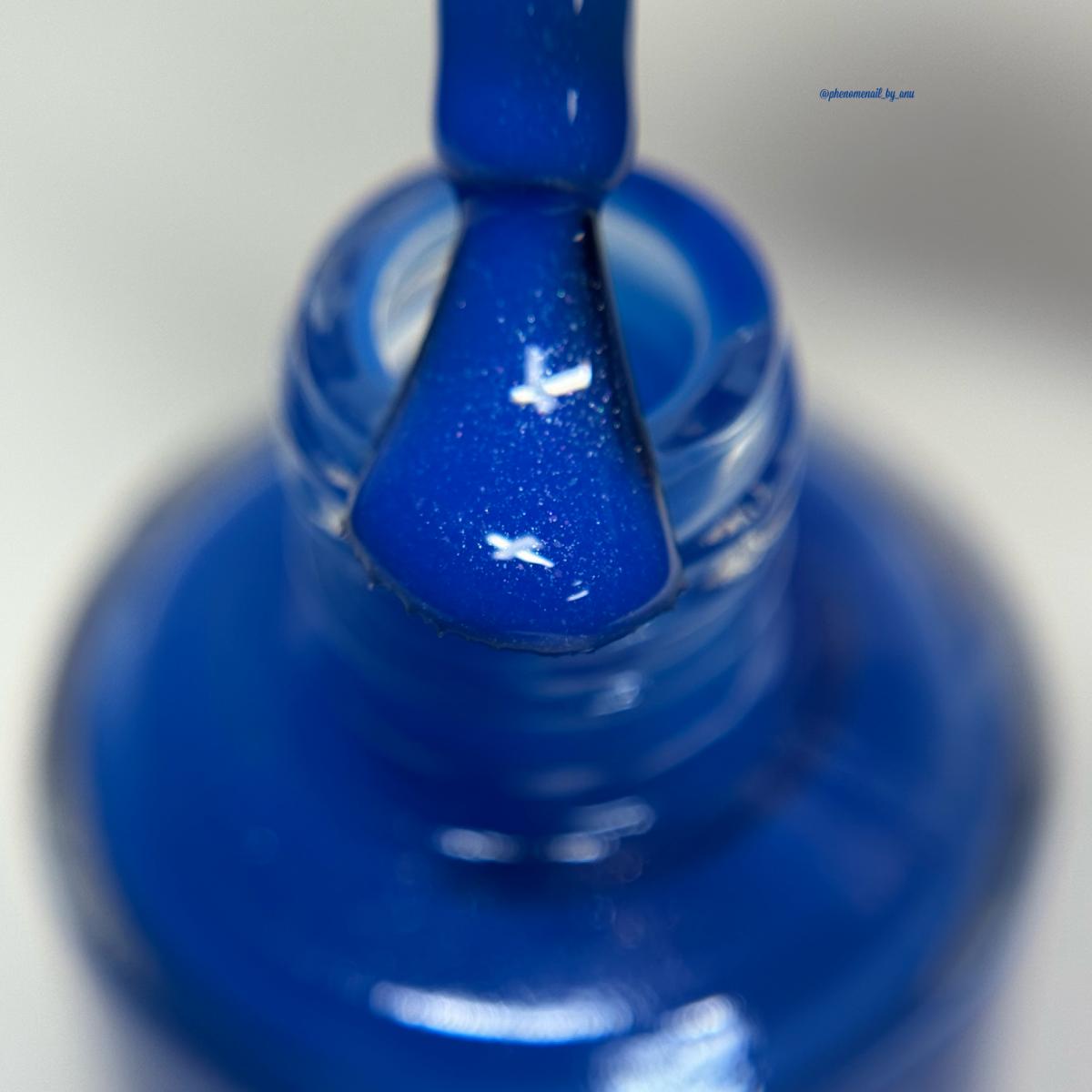 A close-up of a cobalt blue nail polish brush applying polish to a clear glass nail bottle, with a small blue brand text watermark in the upper right.