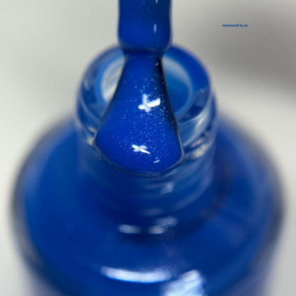 A close-up of a cobalt blue nail polish brush applying polish to a clear glass nail bottle, with a small blue brand text watermark in the upper right.